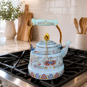 May include: A light blue tea kettle featuring a colorful floral pattern with red, yellow, and green accents. The kettle has a gold-toned metal handle, a matching light blue handle grip, a gold-toned finial on the lid, and a gold-toned chain.