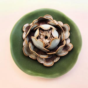 May include: A ceramic decorative piece with a flower design. The flower has layers of brown and light green petals, with a gold-coloured centre. The flower sits on a round, dark green base.