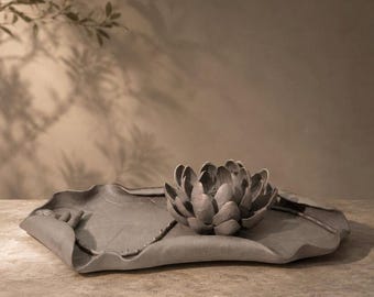May include: A matte grey ceramic decorative tray shaped like a large lily pad with a sculpted lotus flower resting in the centre. The piece features organic, undulating edges and a textured surface that mimics the natural veins of a leaf.