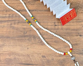 May include: A long necklace featuring white beads, accented with smaller turquoise, red, and yellow beads. Two black and white patterned beads are integrated into the strand. The necklace ends with a silver-toned metal charm and two small dangling bead strands.