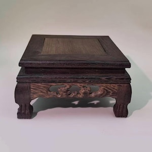 May include: Dark brown wooden display stand with a square top and carved details. The stand has a layered design with decorative cutouts on the lower section and curved legs. The wood grain is visible, adding texture to the piece.