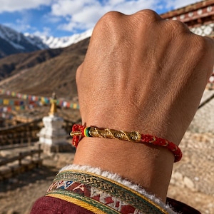 May include: A red cord bracelet featuring a central section of twisted metallic gold thread. The bracelet includes a small decorative accent with thin bands of green and yellow thread near the gold section, secured with a simple knot closure.