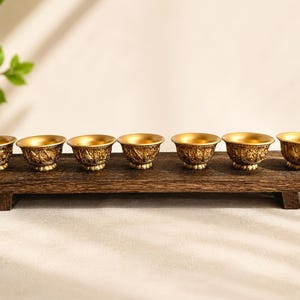 May include: A set of seven small, round, gold-colored metal cups with intricate embossed detailing on the exterior. The cups are arranged in a single horizontal row, resting on a long, rectangular, dark brown wooden tray with short legs.