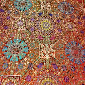 May include: A vibrant red fabric featuring a repeating geometric pattern of interconnected squares and diamonds. The design includes intricate floral medallions in shades of turquoise, yellow, purple, and gold, creating a dense, ornate textile surface.