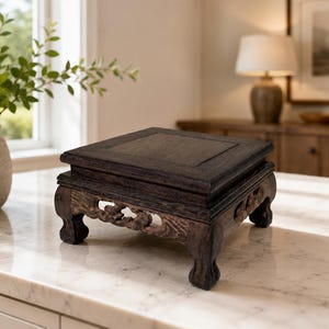 May include: A square, dark brown wooden display stand featuring a flat top surface and four carved legs with intricate openwork detailing. The stand has a visible wood grain texture and a raised border around the perimeter of the top platform.