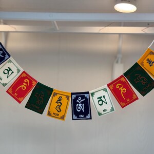 May include: A colorful prayer flag garland with rectangular flags. Each flag features a different color, including blue, white, red, green, and yellow. The flags display Tibetan script in various colors, strung together with a white cord.