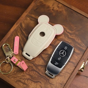 May include: A beige leather key fob cover with pink trim and a matching keychain. A Mercedes-Benz key is next to the cover. The key fob cover has a unique design with two small rounded ears.