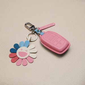 Puede incluir: Una funda rosa de cuero para la llave del coche y un llavero con forma de flor colorida. El llavero tiene un diseño de cara sonriente con pétalos azules, blancos, rosas y rojos. La funda de la llave es rosa y está sujeta a un llavero plateado.