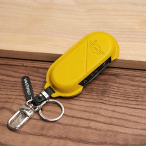 May include: Yellow leather key fob cover with a silver keyring and clasp. The cover has a black accent and a Mini logo. A small black tag with the number "0984525484" is attached.