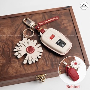 Puede incluir: Una funda de llavero de coche color crema con costuras rojas y un acento rojo, junto a un llavero de margarita blanca con un centro rojo. El llavero tiene el texto "Bao Thien Hai Dang" en una correa roja. Un cierre dorado conecta los artículos.