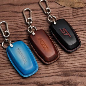 May include: Three leather key fob covers in blue, brown, and black, each with a silver-tone metal clasp. The blue cover has "RAPTOR" embossed, the brown has "F150", and the black has "ST" in red.