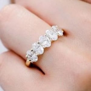 Könnte beinhalten: Ein goldener Ring mit fünf klaren, herzförmigen Edelsteinen. Der Ring wird an einem Finger getragen, wobei die Edelsteine in einer Reihe angeordnet sind. Das Band des Rings ist goldfarben und die Edelsteine funkeln.