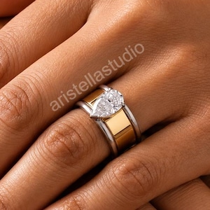 May include: A two-tone ring featuring a pear-shaped clear gemstone set in a polished gold band with silver-toned edges. The ring is worn on a finger, and the text aristellastudio is visible near the centre of the image.
