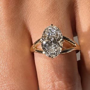 May include: A gold ring featuring a large, oval-cut, clear faceted gemstone set in a prong setting. The band is crafted from polished yellow gold and splits into two separate, slender arms that meet at the base of the central stone setting.