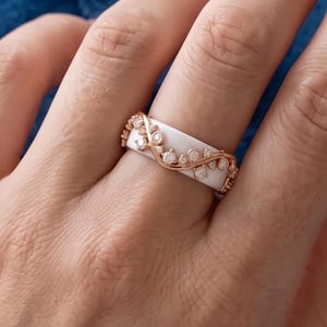 Könnte beinhalten: Ein weißer Keramikring mit einer roségoldfarbenen Metallauflage in Form eines zarten, geschwungenen Rankenmusters. Das Metalldesign ist mit kleinen, klaren, rund geschliffenen Steinen besetzt, die entlang der Kurven eingebettet sind.