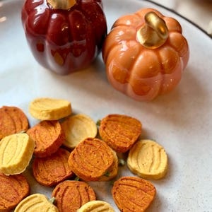 May include: Two glossy ceramic pumpkins, one dark red and one orange, both featuring metallic gold stems, are positioned above a collection of small, flat, pumpkin-shaped cookies in shades of yellow and orange on a speckled white plate.