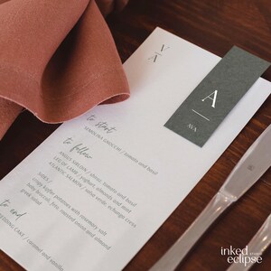 May include: A white wedding menu with a grey place card that says "AVA" on it. The menu lists the food options for the wedding reception, including semolina gnocchi, angus sirloin, leg of lamb, atlantic salmon, and wedding cake.