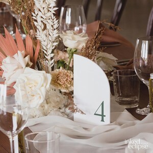 May include: White ceramic table number with a rounded edge and the number "4" in green font. The table number is placed on a white tablecloth with a floral arrangement in the background.