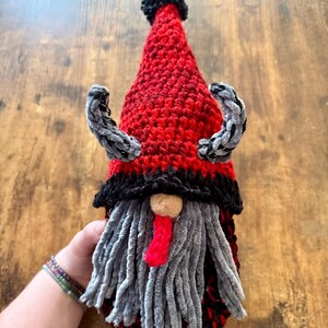 May include: A handmade crochet gnome doll with a red and black hat, gray beard, and small horns. The gnome has a red tongue and a tan nose. The doll is approximately 10 inches tall.