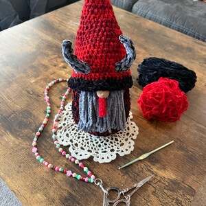 May include: A handmade crochet gnome with a red pointed hat, gray beard, and black accents. A beaded lanyard with scissors, a crochet hook, and yarn are also on the wooden table. The gnome is on a white doily.