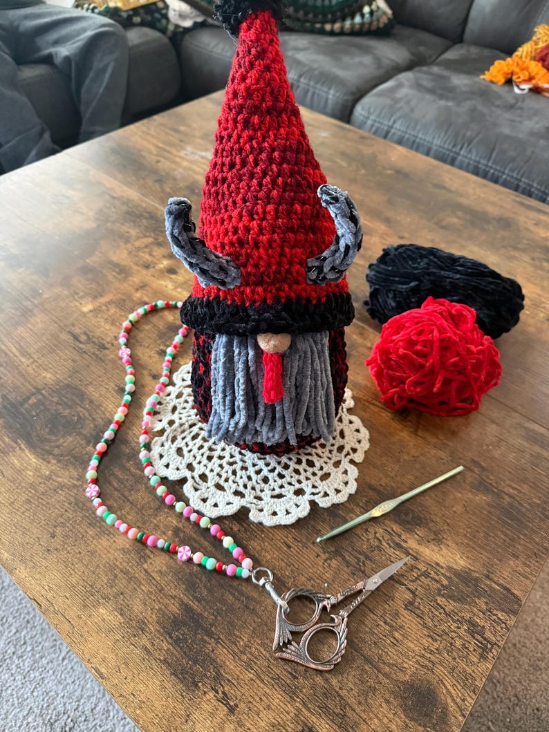 May include: A handmade crochet gnome with a red pointed hat, gray beard, and black accents. A beaded lanyard with scissors, a crochet hook, and yarn are also on the wooden table. The gnome is on a white doily.