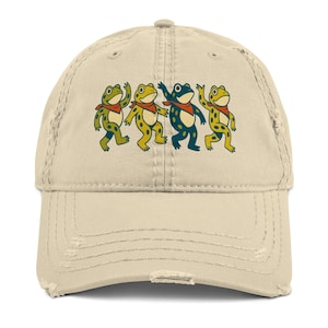 Può includere: Un cappellino da baseball beige invecchiato con una grafica di quattro rane danzanti in verde e giallo, ognuna con una bandana rossa. Il cappellino ha un aspetto usurato con bordi sfilacciati.
