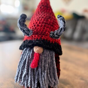 May include: A handmade gnome with a red and black crocheted hat, gray beard, and small gray horns. The gnome has a red tongue and a small tan nose. The gnome is a decorative item.