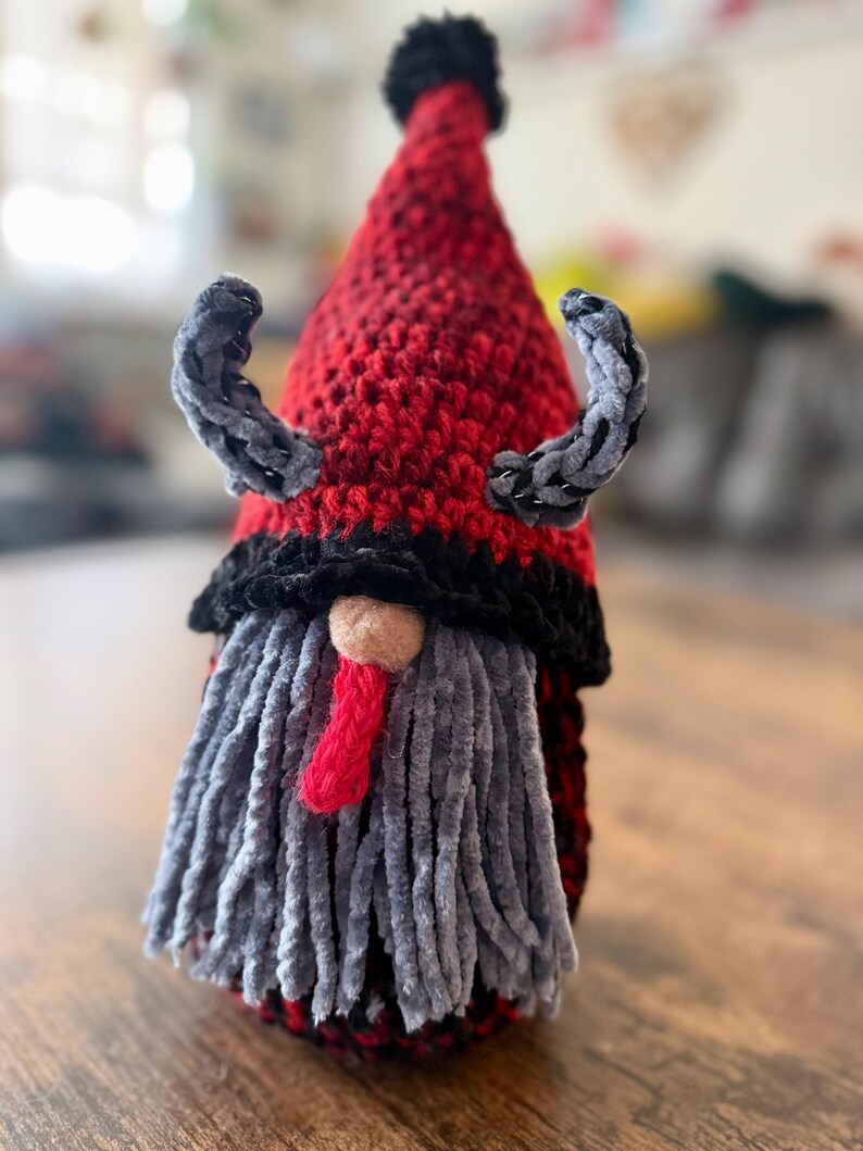 May include: A handmade gnome with a red and black crocheted hat, gray beard, and small gray horns. The gnome has a red tongue and a small tan nose. The gnome is a decorative item.