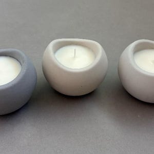 May include: Three spherical candle holders in shades of gray and off-white. Each holder contains a white candle with a central wick. The holders have a smooth, rounded shape, creating a minimalist aesthetic.