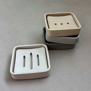 May include: A stack of four square soap dishes in neutral tones. The top dish is a light beige, with three small holes. The other dishes are gray and white. The bottom dish has three vertical slots.