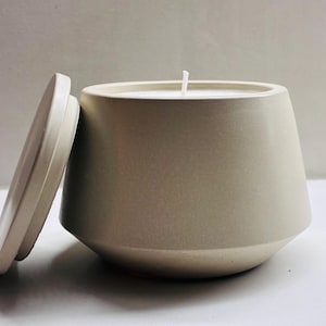Op de afbeelding: Een beige kaars in een moderne, geometrisch gevormde container met een bijpassende deksel. De kaars heeft een witte lont en een gladde, matte afwerking. De deksel staat iets open.