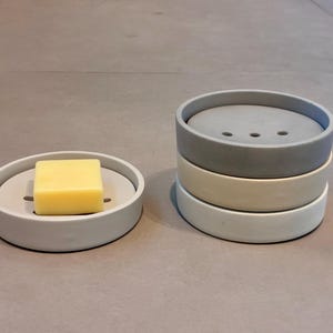 May include: A light gray soap dish with a yellow bar of soap. Stack of three soap dishes in gray, off-white, and white. The soap dish has a circular design with a raised edge and drainage slots.
