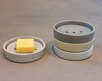 May include: A light grey soap dish with a yellow bar of soap. Stack of three soap dishes in grey, off-white, and white. The soap dish has a circular design with a raised edge and drainage slots.