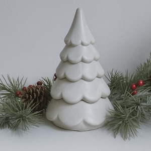 May include: A ceramic Christmas tree in a soft white color, featuring a tiered design with scalloped edges. The tree is surrounded by green pine branches, red berries, and a pine cone, creating a festive holiday scene.
