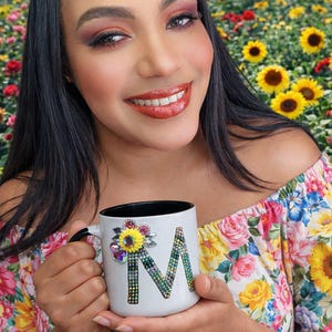 Puede incluir: Una taza de cerámica blanca con un asa negra y el texto "But First, Coffee" en negrita negra. La taza se encuentra sobre un fondo borroso, sugiriendo un ambiente acogedor.