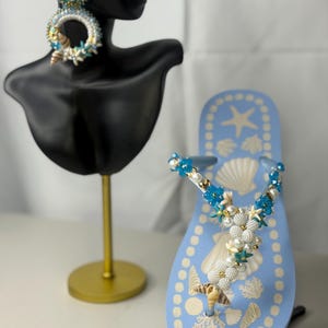 Pode incluir: Uma sandália havaiana azul clara decorada com conchas, pérolas e detalhes florais azuis. A sandália havaiana apresenta um design de concha e é exibida ao lado de um par de brincos de contas com tema de concha.