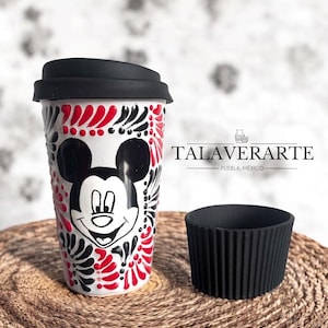 May include: A white ceramic travel mug featuring a Mickey Mouse design with red and black floral accents. The mug has a black lid and a black silicone sleeve. The text "TALAVERARTE - PUEBLA, MEXICO" is printed on the mug.