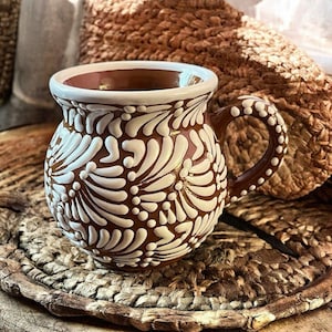 Puede incluir: Una taza de cerámica marrón con un diseño floral blanco. La taza tiene un cuerpo redondeado, un asa y un borde blanco. El patrón floral está en relieve y cubre la mayor parte de la superficie de la taza. La taza está sobre una estera tejida.
