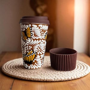 May include: A travel coffee mug with a brown lid and a matching sleeve. The mug features a white, yellow, and brown floral pattern. It sits on a round, woven placemat on a wooden table.