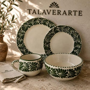 Puede incluir: Un juego de vajilla de cerámica que incluye dos platos redondos, un cuenco y una taza, todos con un esmalte blanco roto y un patrón de plumas de color verde oscuro pintado a mano en los bordes. La palabra TALAVERARTE es visible en la pared.