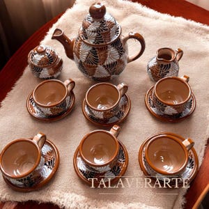 May include: A ceramic tea set featuring a teapot with a lid, a sugar bowl with a lid, a creamer, and six cups with matching saucers. Each piece is decorated with a repeating fan-like pattern in shades of brown, black, and white. The text TALAVERARTE is visible.