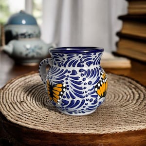 May include: A blue and white ceramic mug with a butterfly design. The mug has a handle and is decorated with yellow and orange butterflies. The mug is sitting on a wooden surface.