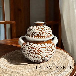 Puede incluir: Una olla de cerámica marrón con tapa, adornada con patrones florales blancos. La olla tiene dos asas y un borde blanco. El texto "TALAVERARTE PUEBLA-MEXICO" es visible en la parte inferior. La olla está sobre un salvamanteles tejido.