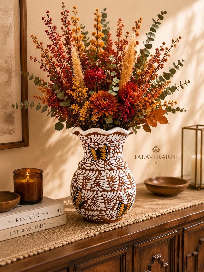 May include: A ceramic vase with a scalloped rim, featuring a brown base with white painted feather-like patterns and butterfly motifs. The vase is filled with a large arrangement of dried flowers in shades of orange, red, and yellow, including eucalyptus.