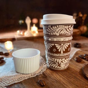 May include: A brown ceramic travel mug with a white lid and a white floral design. The mug is sitting on a wooden table with coffee beans scattered around it.