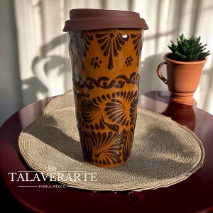 May include: A brown ceramic travel mug with a dark brown lid and intricate floral patterns. The mug features the text "TALAVERARTE - PUEBLA, MÉXICO." The mug is on a round woven placemat.