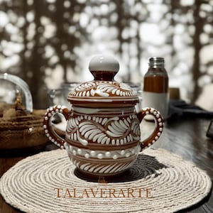 May include: A brown and white ceramic jar with a lid and two handles. The jar is decorated with white floral patterns and a white beaded design. The lid has a white knob. The jar is sitting on a round, woven placemat. The text "TALAVERARTE" is visible.