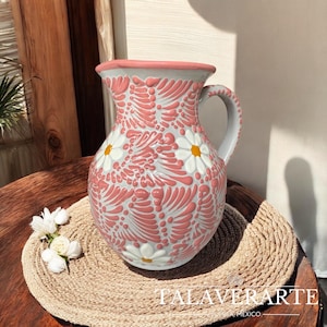 Puede incluir: Jarra de cerámica pintada a mano con diseño floral. La jarra presenta una base blanca con un patrón floral rosa y margaritas blancas con centros amarillos. El borde y el asa son de color gris claro. La jarra está sobre una estera tejida.