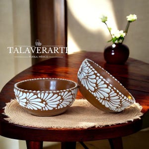Puede incluir: Dos cuencos de cerámica marrón con diseños florales blancos. Los cuencos están sobre una esterilla tejida en una mesa de madera oscura. Los cuencos son de Talaverarti, Puebla, México.