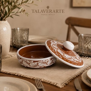 Puede incluir: Un plato redondo de cerámica color terracota con tapa a juego, que presenta intrincados patrones florales blancos pintados a mano. El plato tiene un acabado esmaltado liso y un pomo blanco en la tapa. El texto TALAVERARTE PUEBLA, MEXICO es visible.